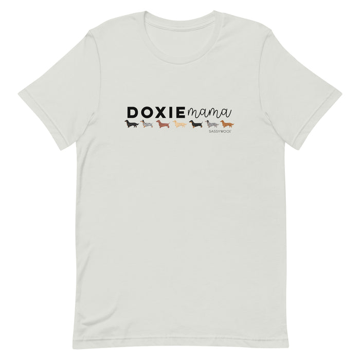 The Doxie Mama Tee by Sassy Woof is a white unisex t-shirt displaying “DOXIE mama” and five colorful dachshund illustrations, with the Sassy Woof logo beneath the last dog—a perfect pick for dachshund lovers.