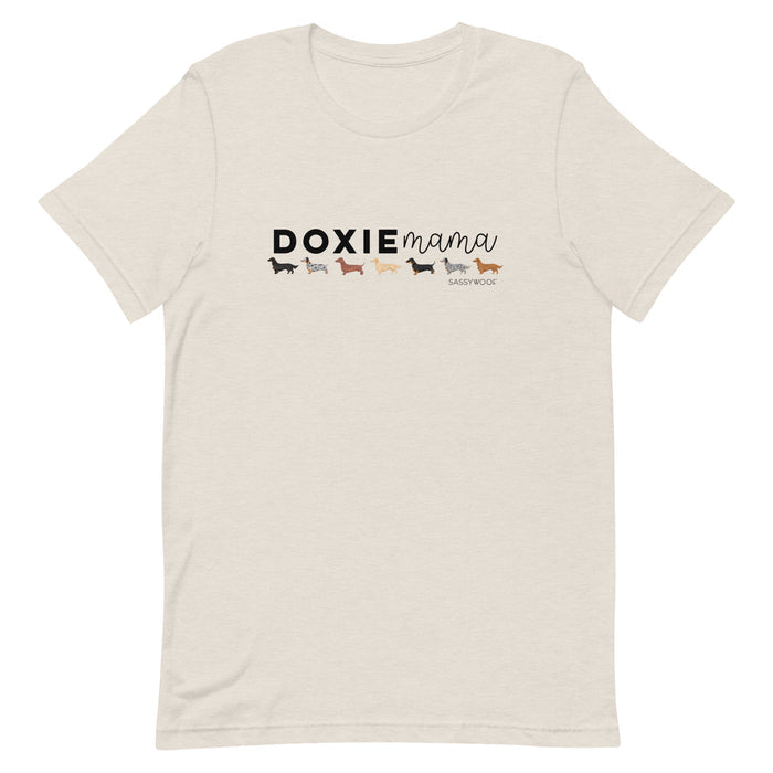 The Doxie Mama Tee by Sassy Woof is a beige unisex t-shirt with “DOXIE mama” and colorful dachshund illustrations—perfect for any dachshund lover.