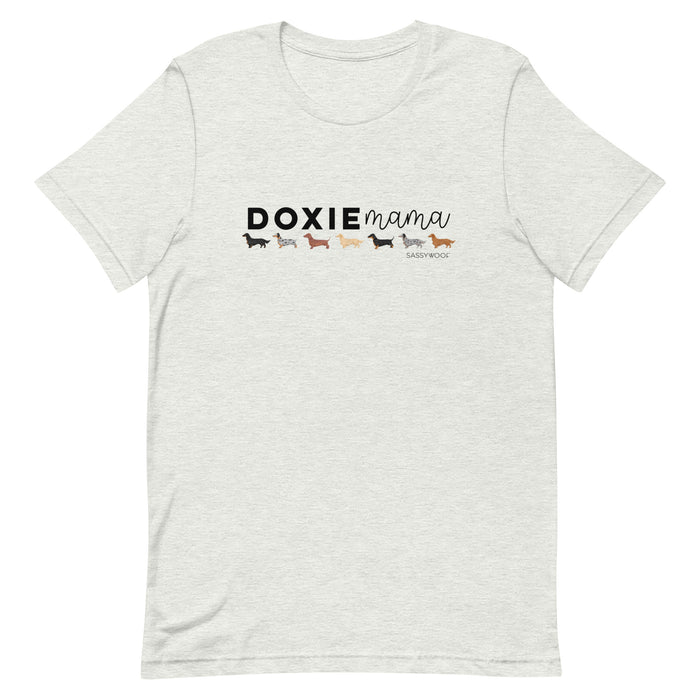 The Doxie Mama Tee by Sassy Woof is a light gray unisex shirt with "DOXIE mama," five colorful dachshunds, and the Sassy Woof logo under "mama." Ideal for dog lovers!.