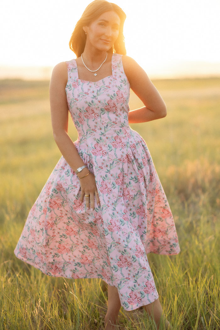 A woman stands in a sunlit field wearing the Edith Dress in Pink Sketched Flower by Ivy City Co, featuring a sweetheart neckline. She smiles gently, with one hand resting on her dress and the other relaxed at her side.