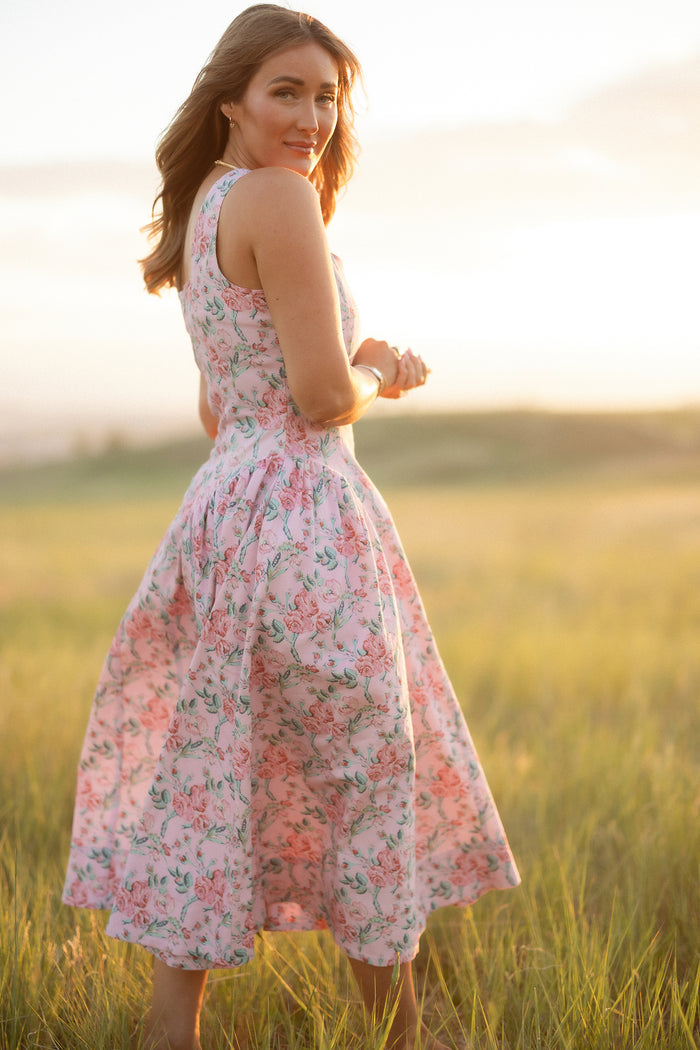 A woman stands in a sunlit field, glancing over her shoulder, wearing the Edith Dress in Pink Sketched Flower by Ivy City Co—a sleeveless dress with a full skirt and sweetheart neckline featuring a delicate floral print.