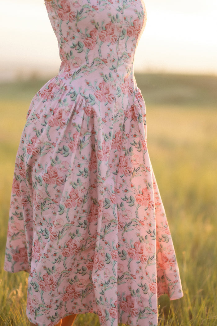A person wears the Ivy City Co Edith Dress in Pink Sketched Flower while standing in a sunlit grassy field. The softly focused, above-the-shoulder crop highlights the dress’s vintage-inspired floral pattern and sweetheart neckline.