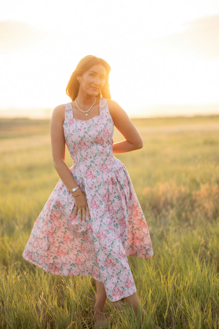 A woman wearing the Edith Dress in Pink Sketched Flower by Ivy City Co stands in a sunlit field, smiling gently as she holds her dress. The soft sunlight creates a warm, glowing effect around her.