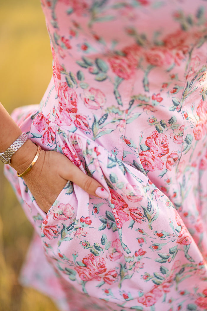 A close-up of a person wearing the Edith Dress in Pink Sketched Flower by Ivy City Co, featuring a sweetheart neckline and pockets. They accessorize with a gold bracelet, watch, and have light pink nails.