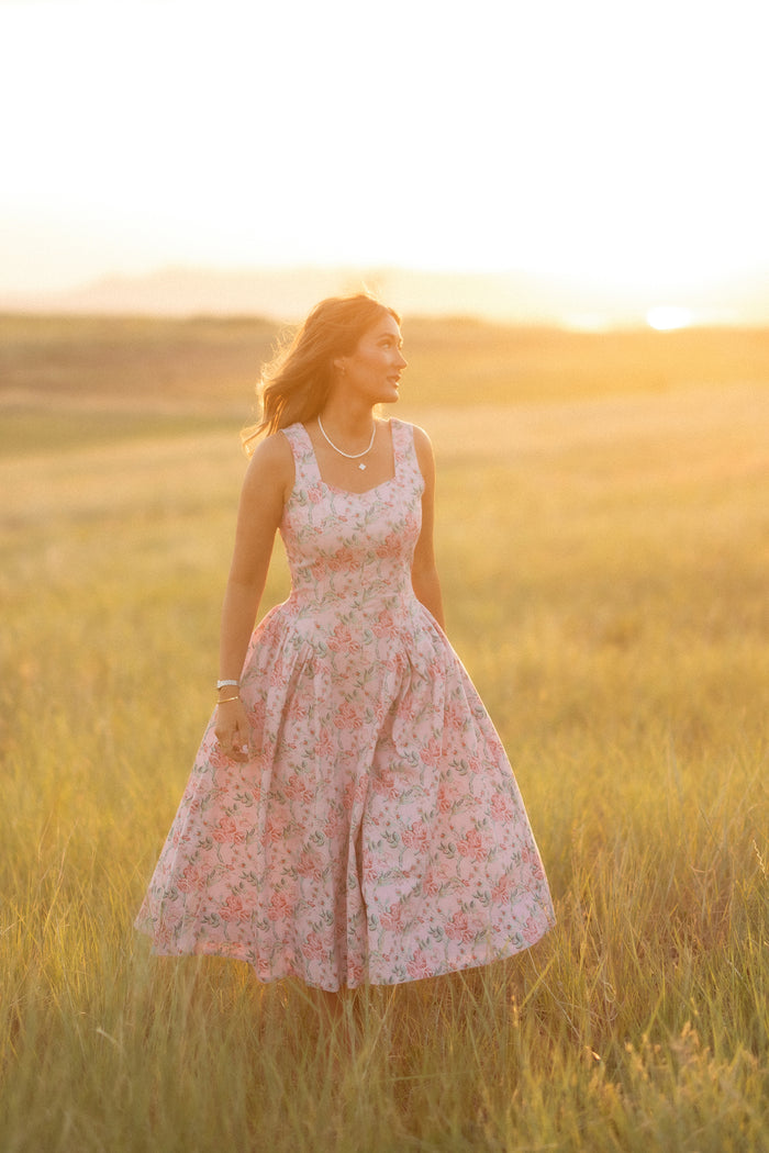 A woman wears the Ivy City Co "Edith Dress in Pink Sketched Flower" as she stands in a sunlit field of tall grass at sunset, looking to the side with a gentle smile as warm light surrounds her.