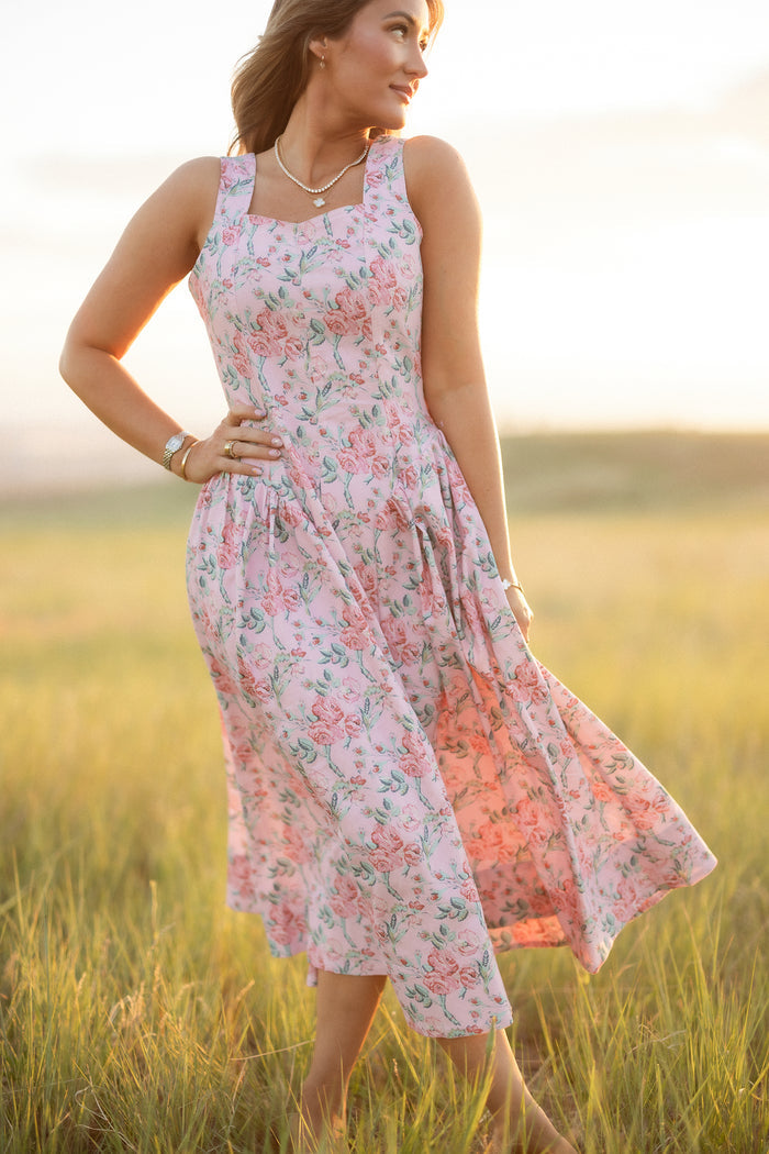 A woman stands in a sunlit meadow wearing the Edith Dress in Pink Sketched Flower by Ivy City Co, holding her skirt gently, with grass and a blurred landscape in the background.