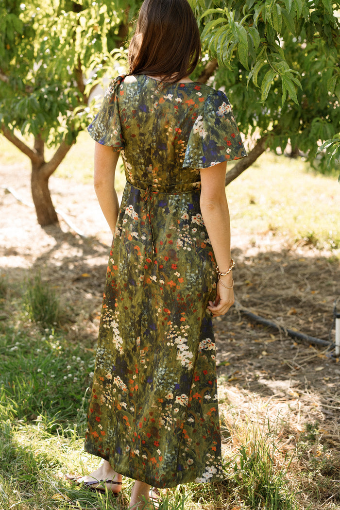 A woman stands outdoors facing away, surrounded by sunlit trees and grass, wearing the Ivy City Co Fiona Flutter-Sleeve Maxi Dress in Impressionist Bloom—a long satin dress adorned with flutter sleeves and a vibrant floral print.