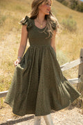 A woman stands outdoors on a dirt path near a wooden fence, holding the skirt of her green floral Hattie Dress in Woodland Floral by Ivy City Co. With long wavy hair and white boots, she poses against a backdrop of grassy hills. This dress features a smocked bodice, is maternity friendly, and perfect for any season.