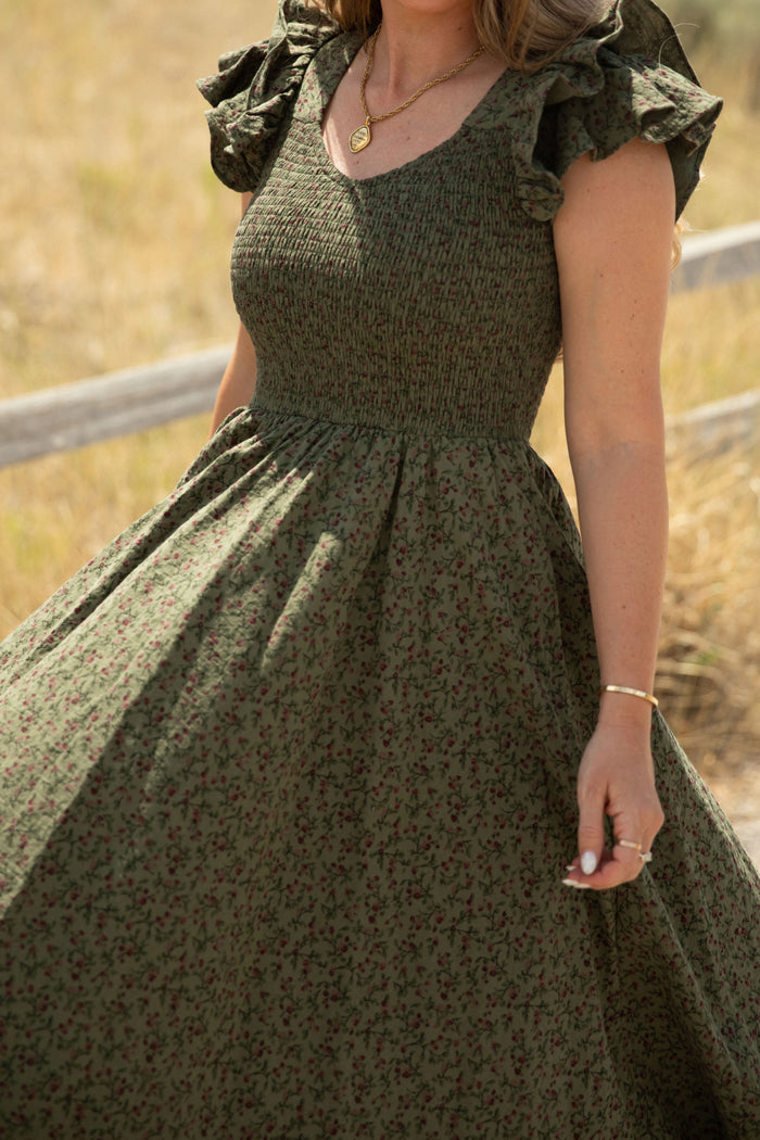 A woman stands outdoors in the Ivy City Co Hattie Dress in Woodland Floral, featuring ruffled sleeves and a smocked bodice. She accessorizes with a gold necklace and bracelet while holding the skirt, with a blurred wooden fence and dry grass in the background. This maternity-friendly style effortlessly combines comfort and elegance.
