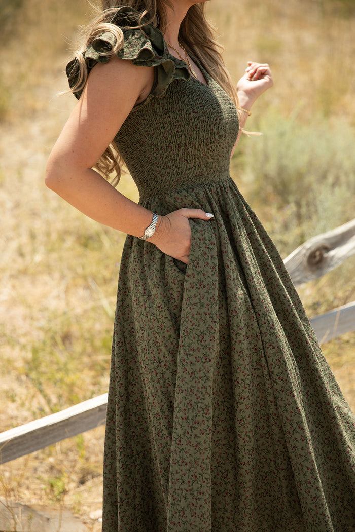 A woman stands outdoors in a sunlit, grassy field with a wooden fence, wearing the Ivy City Co Hattie Dress in Woodland Floral. The green dress features ruffled sleeves, a smocked bodice, and practical pockets—highlighting its breezy, maternity-friendly design.