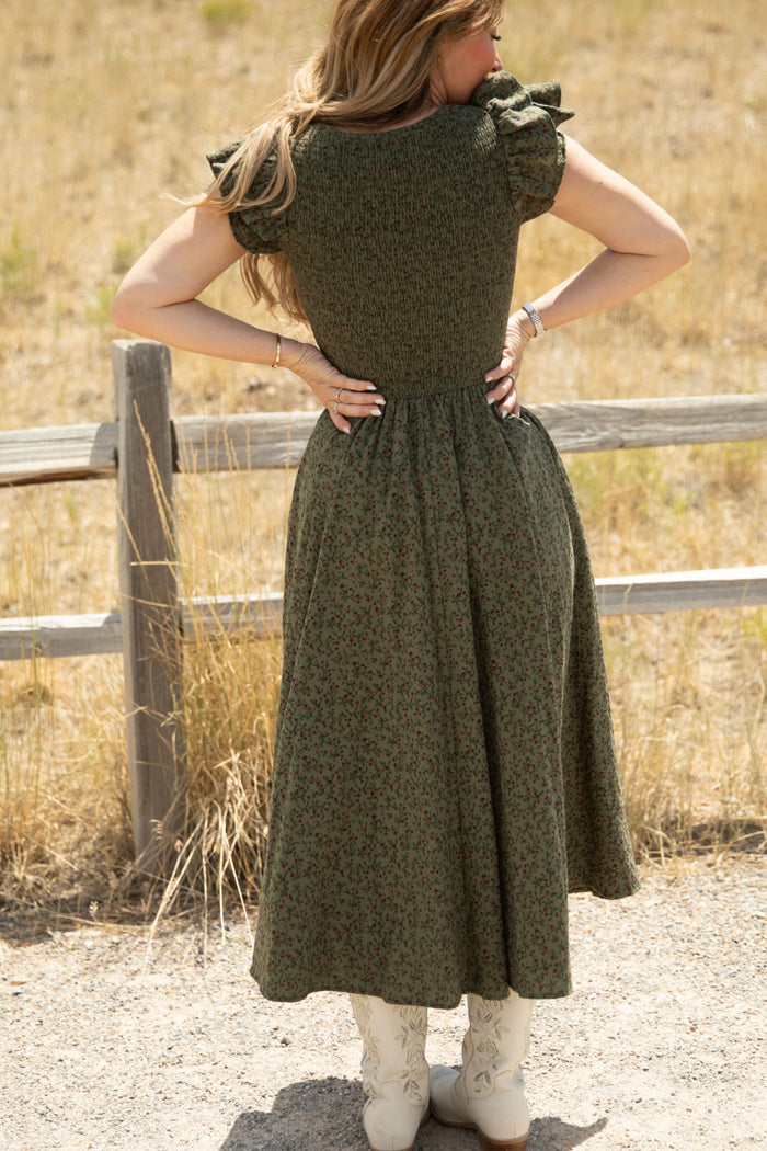 Outdoors by a wooden fence, a woman stands with her hands on her lower back, facing away and wearing the Ivy City Co Hattie Dress in Woodland Floral—a green floral dress featuring a smocked bodice and ruffled sleeves—paired with white cowboy boots. Maternity friendly and effortlessly stylish, she is framed by dry grass and a dirt path.