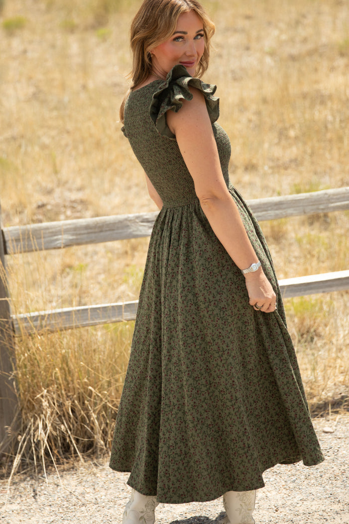 Outdoors amid dry grass and a wooden fence, a woman faces slightly away, looking over her shoulder as she wears the Ivy City Co Hattie Dress in Woodland Floral—a long, green floral dress featuring ruffled sleeves and a smocked bodice—styled with white boots and a watch. This maternity-friendly design from Ivy City Co effortlessly blends comfort with timeless elegance.