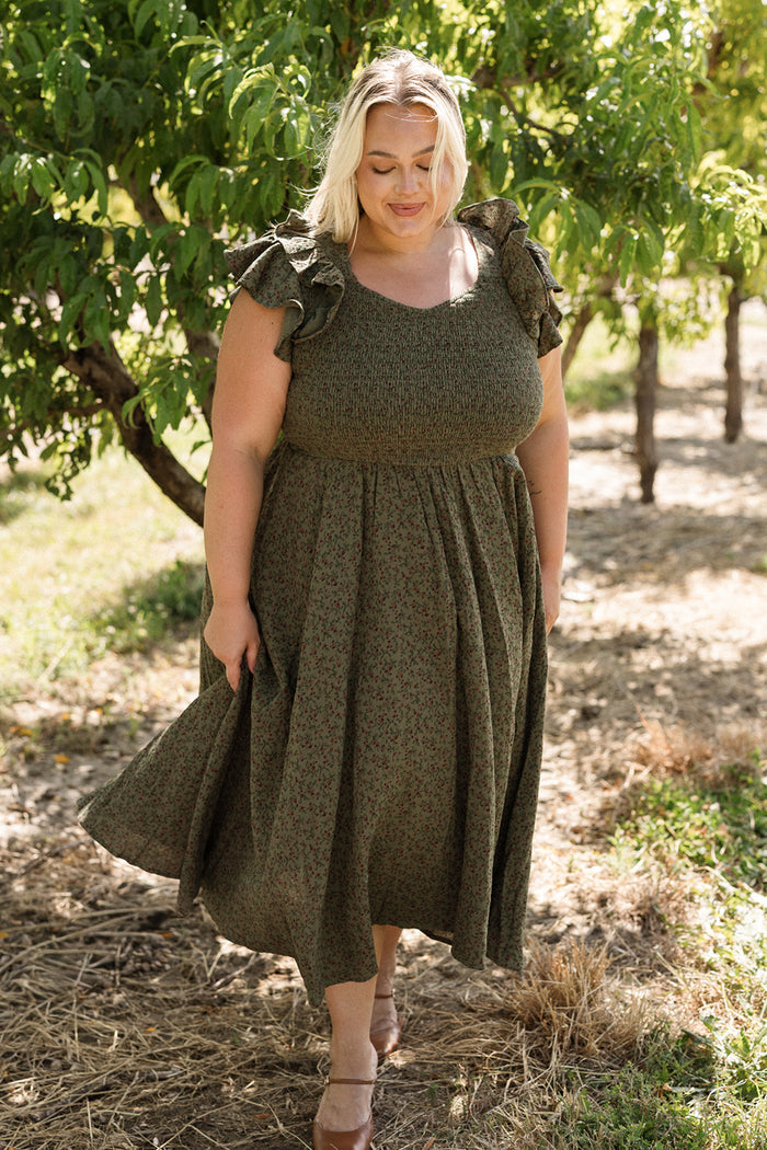 A woman wearing the Ivy City Co Hattie Dress in Woodland Floral, featuring a smocked bodice and a maternity-friendly design, strolls outdoors on a sunny day amidst trees and grass, smiling and looking down as she radiates comfort and charm.