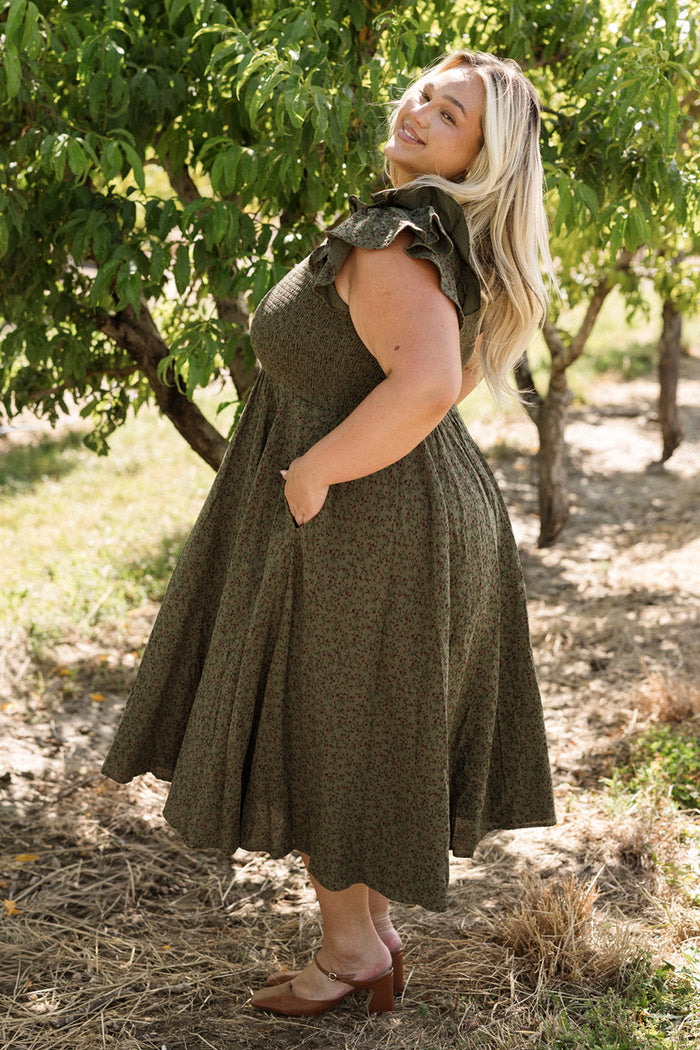 A woman with long blonde hair stands outdoors near leafy trees, smiling with one hand in her pocket as sunlight filters through the branches. She wears the Ivy City Co Hattie Dress in Woodland Floral—a green, ruffled sleeveless dress with a smocked bodice and a maternity-friendly fit—paired with brown heels.