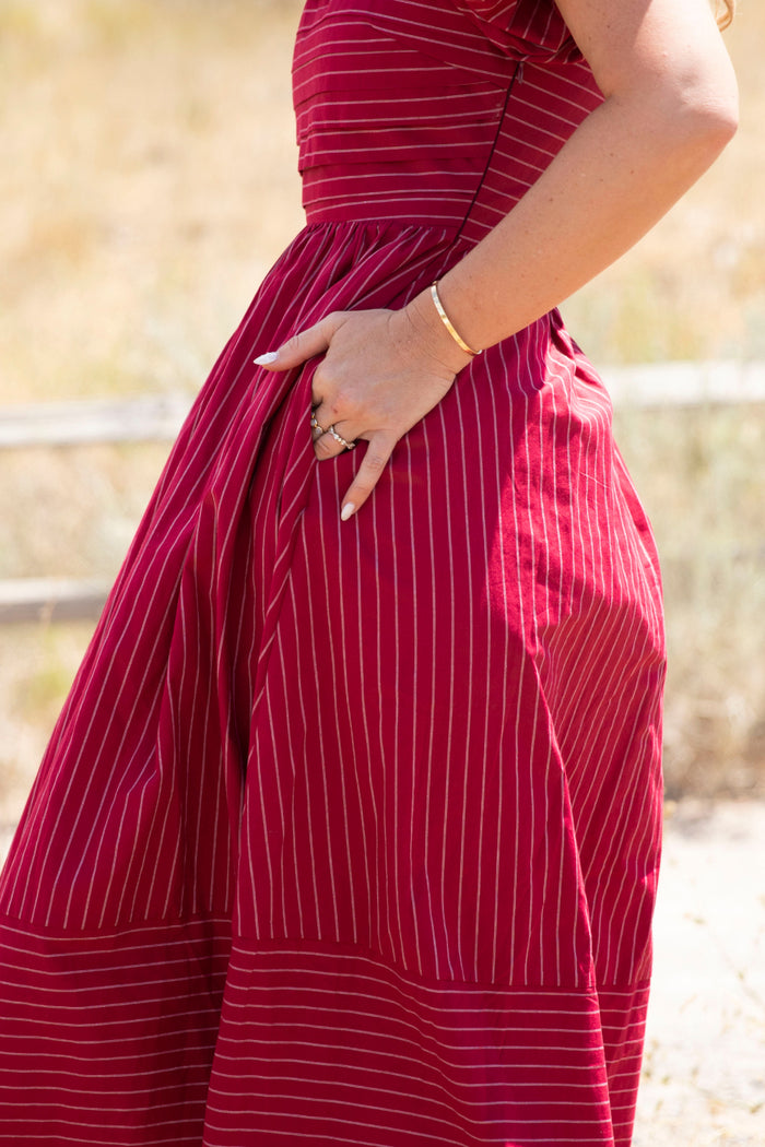 A person stands outdoors wearing the Julie Dress in Cranberry Pinstripe by Ivy City Co, featuring white vertical stripes, puff sleeves, and a smocked bodice. Their right hand is tucked into the dress pocket, displaying a bracelet and rings. The sunlit background is softly blurred.