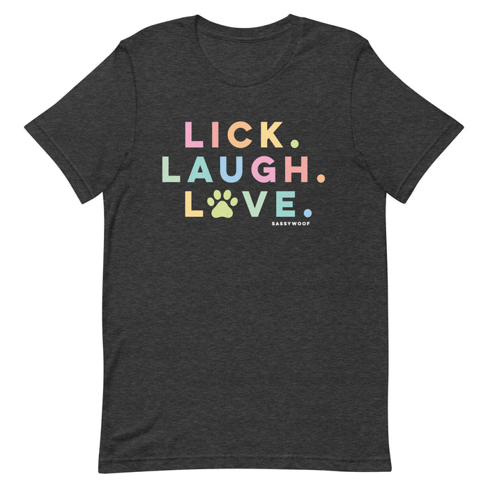 The Lick, Laugh, Love Tee by Sassy Woof is a dark gray, lightweight unisex pet shirt featuring colorful text—LICK. LAUGH. LOVE.—with a paw print for the O and the brand name below the design.
