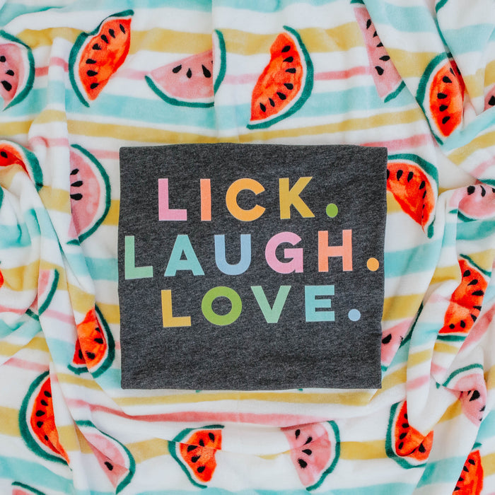 The Sassy Woof Lick, Laugh, Love Tee is a lightweight summer dog t-shirt with colorful lettering, displayed on a blanket with watermelon slices and pastel stripes.