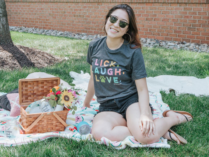 A smiling woman in sunglasses sits on a blanket by a picnic basket, wearing the Sassy Woof Lick, Laugh, Love Tee and denim shorts. Flowers, snacks, and a brick wall appear in the background.
