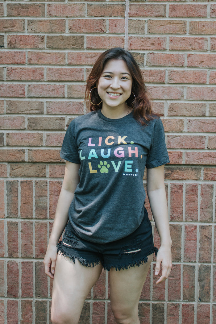 A young woman smiles in front of a brick wall, wearing Sassy Woof’s Lick, Laugh, Love Tee and black denim shorts. The dark, lightweight shirt features “Lick. Laugh. Love.” in colorful letters.