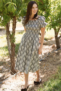 A woman stands outdoors on the grass near trees, smiling and looking to the side, sunlight filtering through the leaves casting dappled light over this maternity-friendly scene. She wears the Ivy City Co Lisa Dress in Jolie Fleur, a floral dress featuring puff sleeves and a smocked bodice.