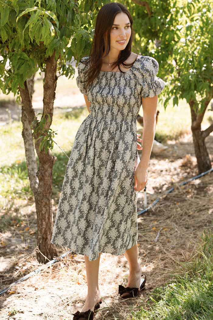 A woman stands outdoors on the grass near trees, smiling and looking to the side, sunlight filtering through the leaves casting dappled light over this maternity-friendly scene. She wears the Ivy City Co Lisa Dress in Jolie Fleur, a floral dress featuring puff sleeves and a smocked bodice.