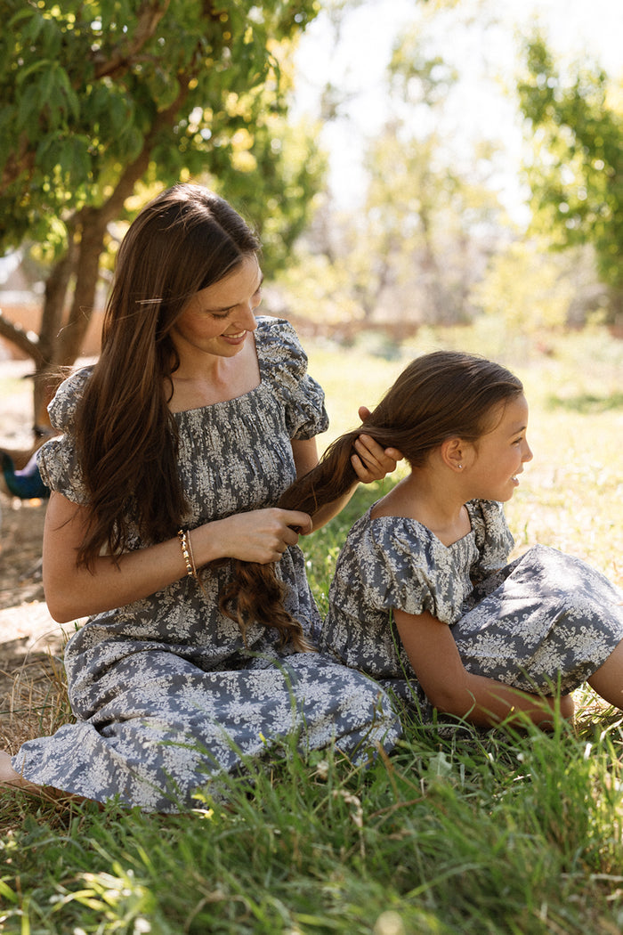 A woman sits on the grass braiding a young girl's hair; both smile beneath a sunlit tree in matching Lisa Dresses in Jolie Fleur by Ivy City Co, featuring smocked bodices that make their outfits perfectly maternity friendly.
