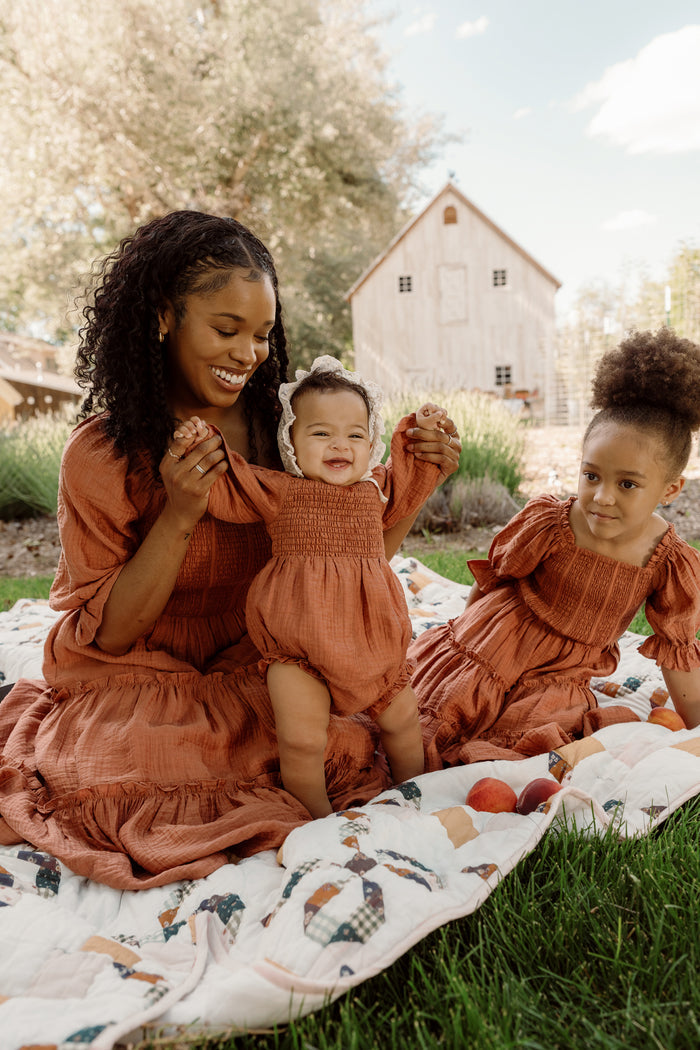 A smiling woman sits on a quilt outdoors, holding a baby’s hands as another child sits beside them. All three wear matching Ivy City Co Madeline Dresses in the warm Apple Cider hue, featuring smocked bodices, with a barn and trees visible in the background.