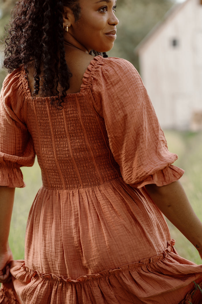 A woman in the Ivy City Co Madeline Dress in Apple Cider, featuring a textured fabric and smocked bodice, stands outdoors looking over her shoulder with a gentle smile. The background is blurred with greenery and a white building—an effortlessly chic and maternity-friendly look.