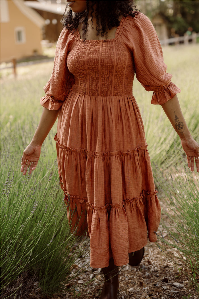 A person wearing the Ivy City Co Madeline Dress in Apple Cider, featuring a long, flowy silhouette and smocked bodice, walks through tall grass with arms slightly outstretched. Blurred greenery and a house in the background showcase the dress’s maternity-friendly design.