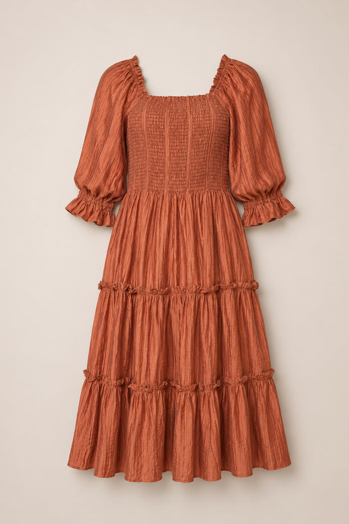 The Madeline Dress in Apple Cider by Ivy City Co features a rust-colored, knee-length silhouette with puffed three-quarter sleeves, a maternity-friendly smocked bodice, and a tiered, ruffled skirt, shown against a neutral background.
