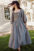 A person stands barefoot on grass, holding the skirt of the Mae 3/4 Sleeves Dress in Prussian Blue Plaid by Ivy City Co, featuring a smocked bodice. Their hair is blowing across their face, and a white building with windows appears in the background.