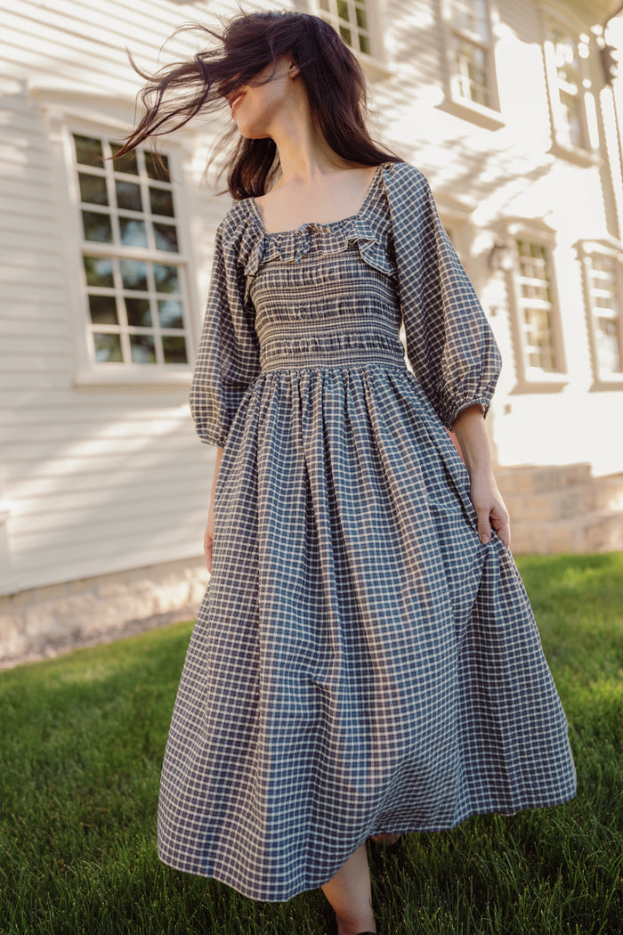 A person stands barefoot on grass, holding the skirt of the Mae 3/4 Sleeves Dress in Prussian Blue Plaid by Ivy City Co, featuring a smocked bodice. Their hair is blowing across their face, and a white building with windows appears in the background.
