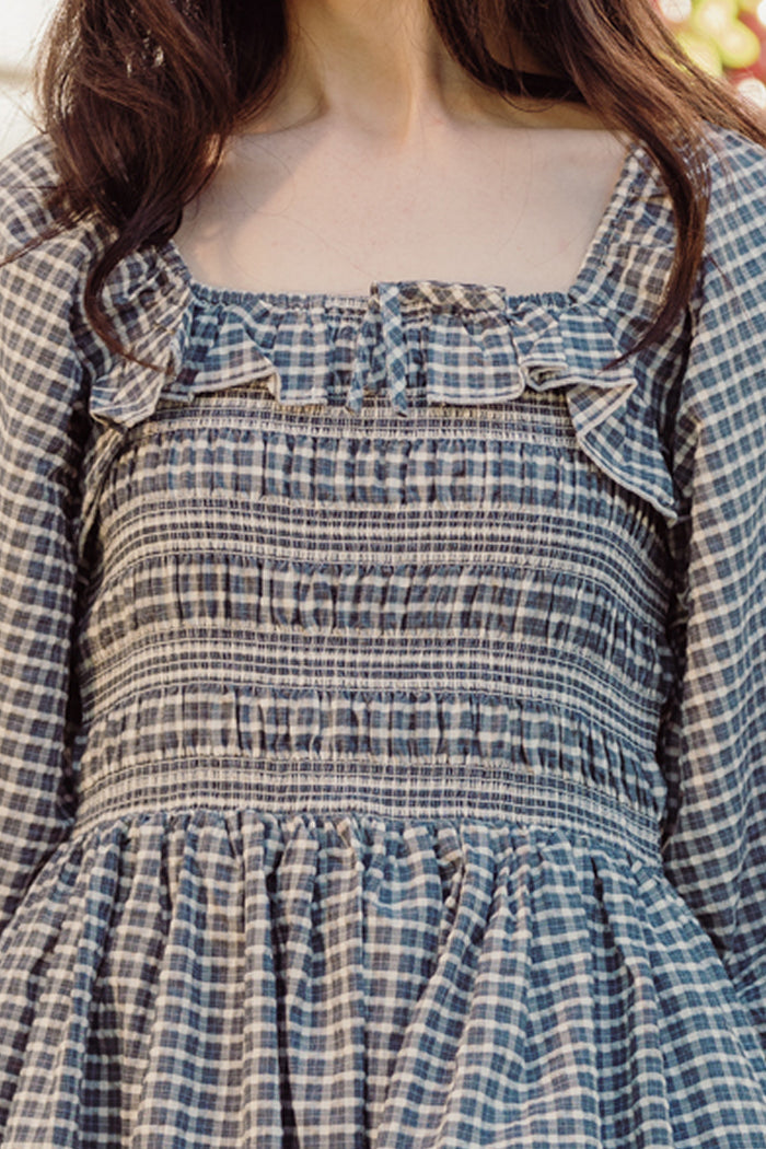 A person standing outdoors wears the Ivy City Co Mae 3/4 Sleeves Dress in Prussian Blue Plaid, which features ruffle accents, a smocked bodice, and a maternity-friendly fit for comfortable yet charming style.