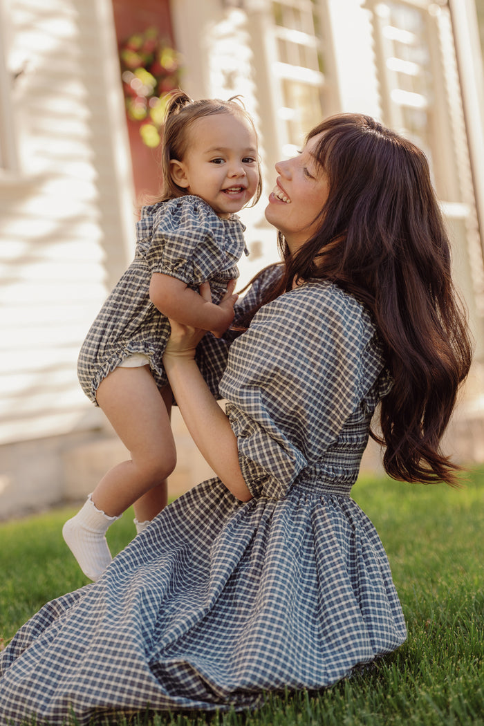 A woman and a toddler, both wearing Ivy City Co's Mae 3/4 Sleeves Dress in Prussian Blue Plaid with smocked bodices, smile and play together on the grass outside a house on a sunny day. The coordinating dresses are maternity friendly, offering added comfort and style.