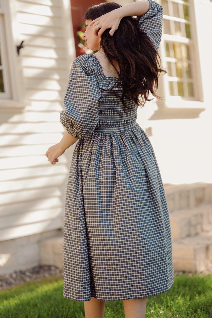 A woman wearing the Mae 3/4 Sleeves Dress in Prussian Blue Plaid by Ivy City Co stands outside a white house, facing away with one hand in her hair and the other by her side as sunlight casts shadows on the wall.