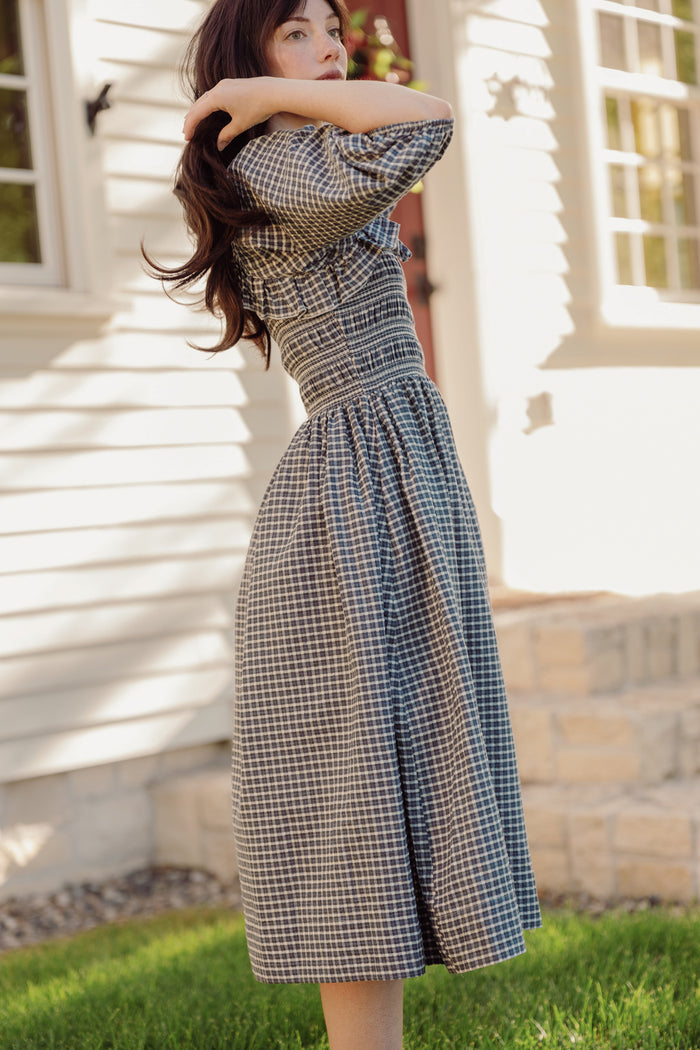 A woman wearing the Mae 3/4 Sleeves Dress in Prussian Blue Plaid by Ivy City Co stands outside on green grass, one arm raised and her hair falling across her face, in front of a white house with stone accents as sunlight casts soft shadows.