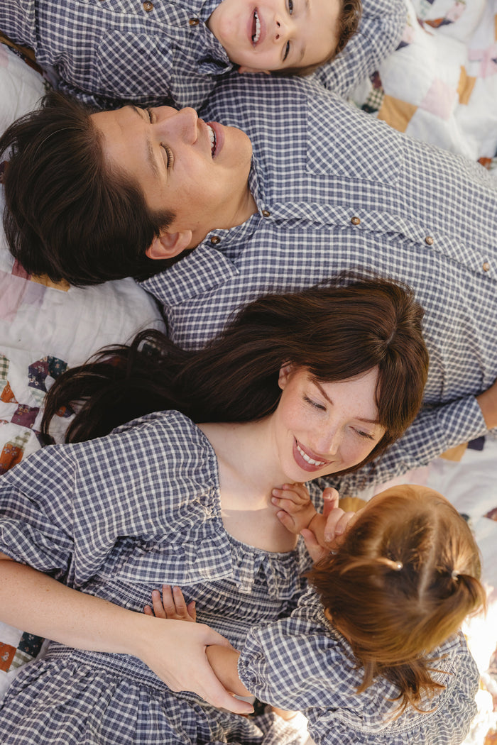 A family of four lies on a blanket, all dressed in matching checkered outfits. The parents smile up at their young children, who beam back and interact playfully. The mother wears the Mae 3/4 Sleeves Dress in Prussian Blue Plaid by Ivy City Co, featuring a comfortable smocked bodice that makes it both stylish and maternity friendly—perfect for relaxed family moments like these.