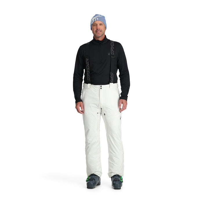 A man standing and wearing white ski pants with adjustable suspenders, a black shirt, and ski boots.