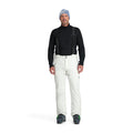 A man standing and wearing white ski pants with adjustable suspenders, a black shirt, and ski boots.