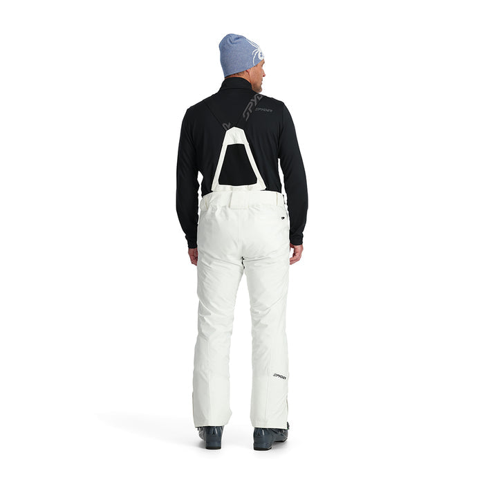 A man standing and wearing white ski pants with adjustable suspenders, a black shirt, and ski boots.