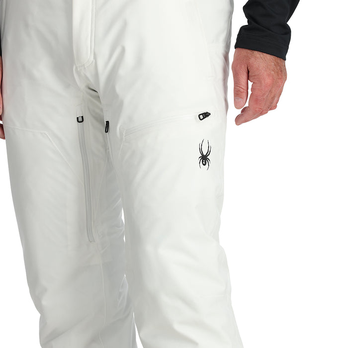 A man standing and wearing white ski pants with adjustable suspenders, a black shirt, and ski boots.