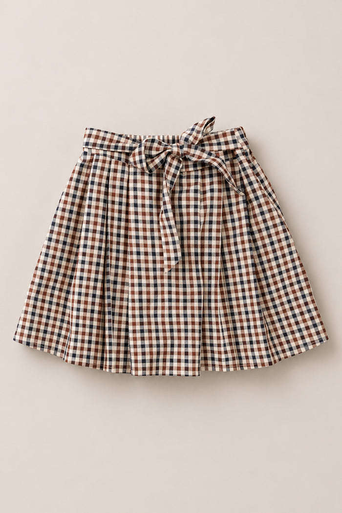 The Mini Addison Skort in Chestnut Gingham by Ivy City Co features a knee-length, pleated silhouette crafted from soft cotton with a brown, white, and blue gingham pattern. Complete with a matching fabric belt tied in a bow at the waist and built-in shorts for added comfort and confidence, it is styled after the signature Mini Addison Skort and is presented laid flat on a light background.