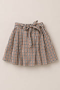 The Mini Addison Skort in Chestnut Gingham by Ivy City Co features a knee-length, pleated silhouette crafted from soft cotton with a brown, white, and blue gingham pattern. Complete with a matching fabric belt tied in a bow at the waist and built-in shorts for added comfort and confidence, it is styled after the signature Mini Addison Skort and is presented laid flat on a light background.