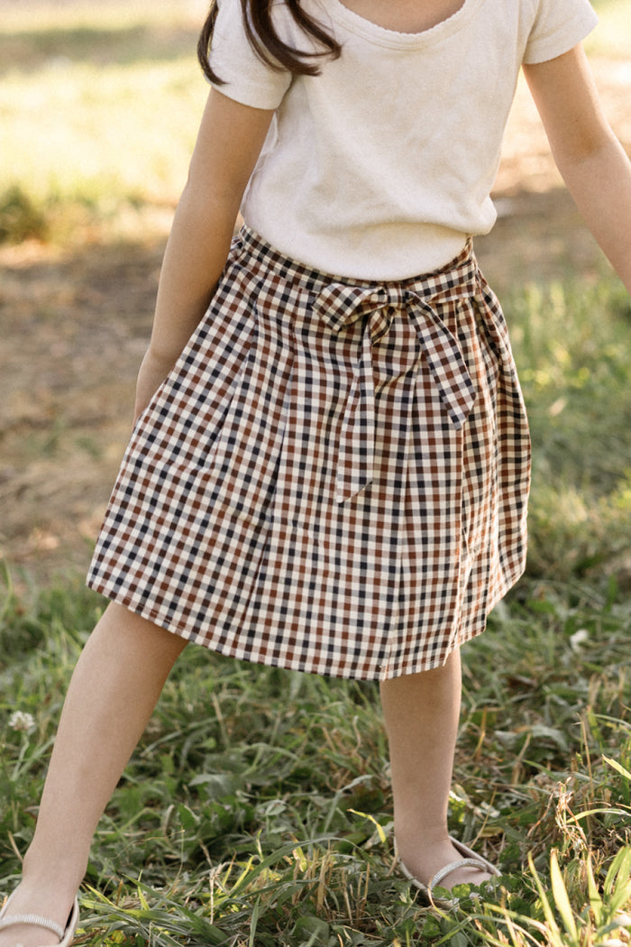 A girl wearing a white t-shirt and the Ivy City Co Mini Addison Skort in Chestnut Gingham, featuring brown, black, and white gingham pattern with built-in shorts and a waist tie, stands in a grassy field.