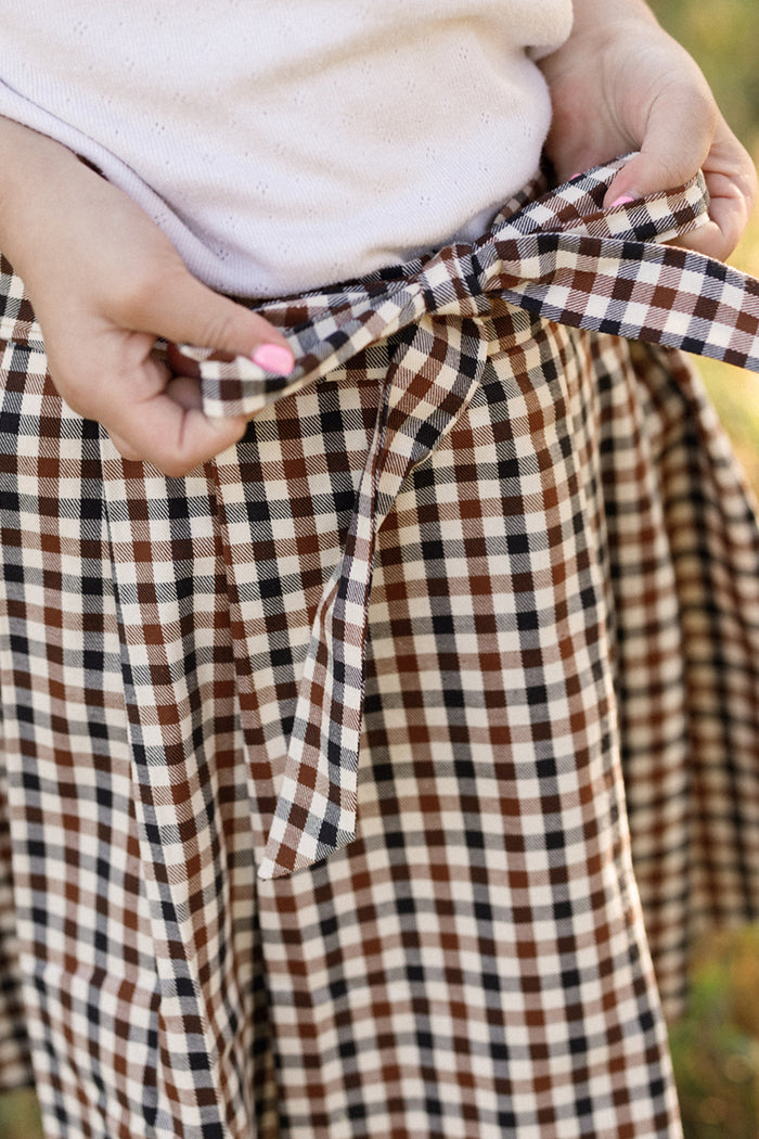 A person ties the bow of an Ivy City Co Mini Addison Skort in Chestnut Gingham, crafted from soft cotton, while wearing a light-colored top. Only their hands and waist are visible.