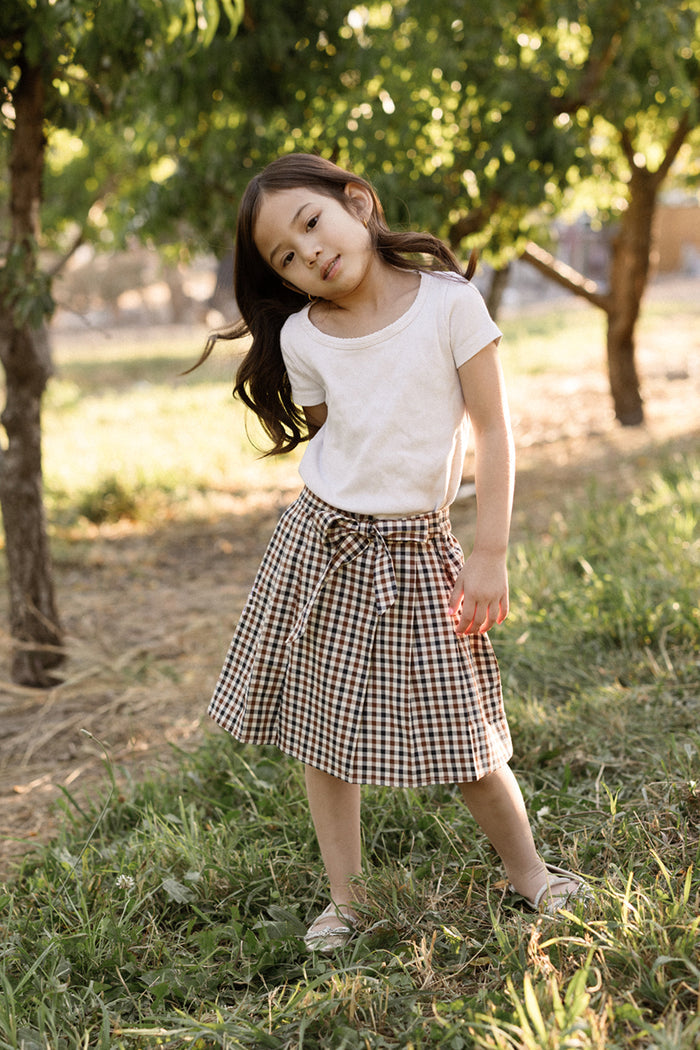 A young girl stands on grass in a sunny outdoor setting with trees in the background, tilting her head and looking at the camera. She is wearing a white t-shirt paired with the Ivy City Co Mini Addison Skort in Chestnut Gingham, featuring soft cotton fabric and built-in shorts.