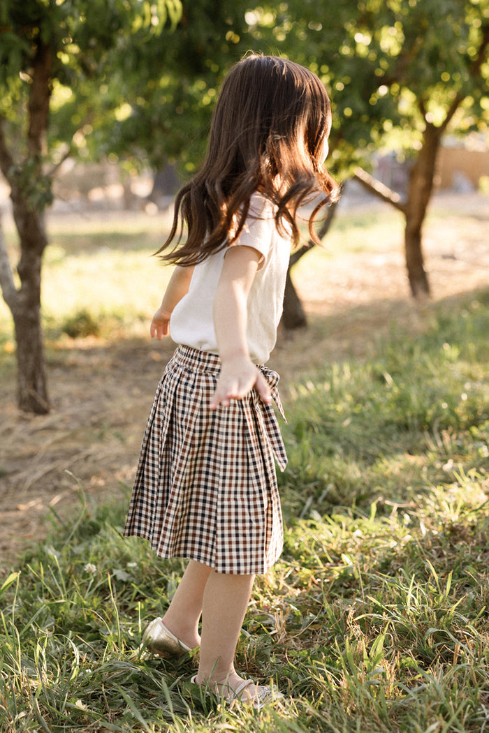 A young girl with long brown hair, dressed in a soft cotton white shirt and the Ivy City Co Mini Addison Skort in Chestnut Gingham with built-in shorts, stands outdoors on the grass with arms outstretched, surrounded by green trees in warm sunlight.