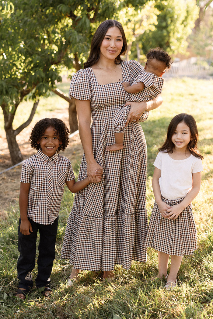 A woman stands outside with three children, all smiling in a sunlit grassy area. The woman, baby, and two older children wear coordinated beige and brown gingham outfits crafted from soft cotton. The girl pairs a white top with the Ivy City Co Mini Addison Skort in Chestnut Gingham, which features built-in shorts for extra comfort.