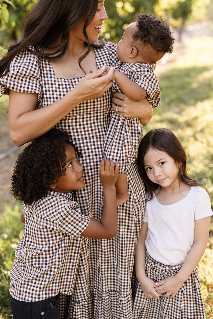 A woman holds a baby while two children stand beside her outdoors. All are dressed in brown plaid patterns, except the girl on the right, who wears a white top paired with Ivy City Co’s Mini Addison Skort in Chestnut Gingham, made from soft cotton. They are smiling and appear happy.