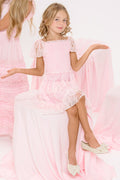 A young girl in the Ivy City Co Mini Addy Dress in Light Pink sits on a draped table, smiling softly. Another person in a similar midi dress stands beside her, mostly out of frame. The scene features light, pastel tones.
